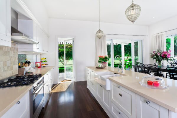 A wide-angle, hyper-realistic, and brightly lit view of a modern kitchen with a minimalist interior design. The room features white cabinets, light-colored stone countertops, a large island with a farmhouse sink, and stainless steel appliances. A set of open patio doors in the background shows a well-manicured yard.