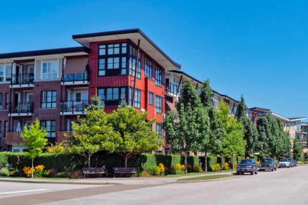 A vibrant, sunny view of a modern residential condominium building with multi-colored siding, including red and white panels, large windows, and balconies. The property is well-maintained with green trees, manicured hedges, and benches along the street.