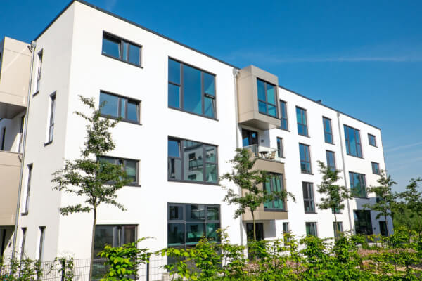 A modern, white residential condominium building with large square windows and glass balconies under a clear blue sky. The exterior is sleek and minimalist, with small trees and green bushes in the foreground.