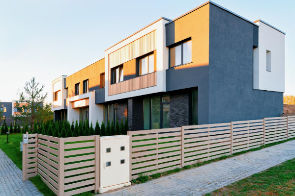 A modern residential complex with multi-story townhouses featuring a blend of white and dark gray facades, accented with wooden slats. A low, beige fence encloses a small yard with green landscaping.
