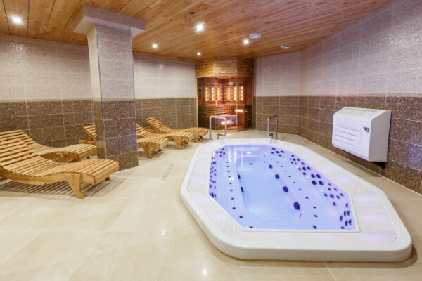 A view of a modern spa and wellness center with a glowing hexagonal jacuzzi in the foreground, wooden lounge chairs, and a glass-enclosed sauna room in the background.