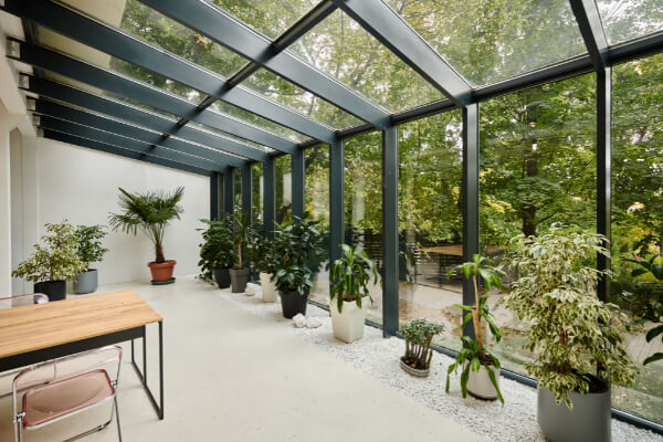 A modern, light-filled sunroom with a glass roof and large windows, offering a panoramic view of the outdoors. The interior features a small wooden desk with a chair, several potted plants, and a white gravel floor, seamlessly blending indoor and outdoor living.