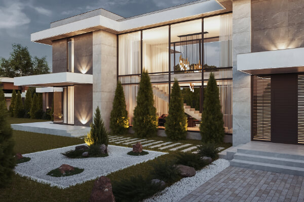 A striking view of a luxurious, modern residential villa at dusk with its interior lights on. The house features a blend of gray stone and white stucco, large floor-to-ceiling windows, and elegant landscaping with white gravel, rocks, and small cypress trees.