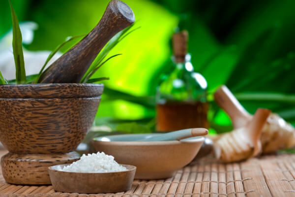 A close-up of natural spa ingredients on a bamboo mat, including a mortar and pestle, sea salt, a small bowl, an oil bottle, and fresh ginger root, set against a blurred green background.