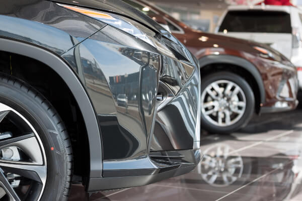 A close-up, low-angle shot of the front bumper and wheel of a glossy black SUV on display inside a car dealership showroom, with another vehicle visible in the background. The polished floor reflects the vehicles and the lights above.