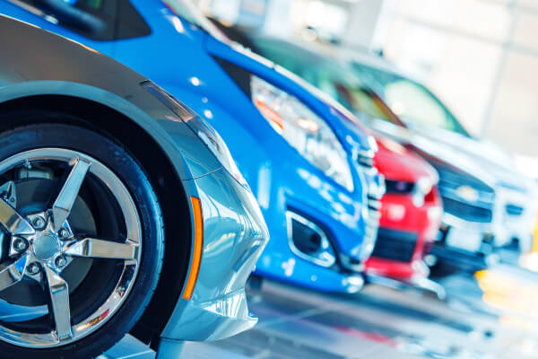 A close-up, low-angle shot of the front ends and wheels of a row of new cars on a polished showroom floor. The vehicles are in various colors, including silver, blue, and red, and are lined up in a row.