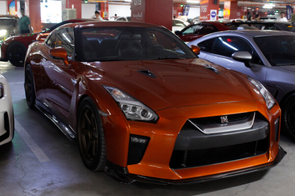 A front-angle, close-up view of a parked Nissan GT-R R35 sports car with a glossy orange finish, black hood vents, and sleek front bumper, surrounded by other cars in an underground parking garage.