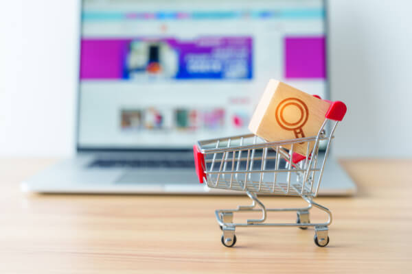 A close-up, eye-level shot of a miniature wooden block with a magnifying glass icon on it, sitting inside a tiny metal shopping cart. In the blurry background, a laptop screen displays an e-commerce website.