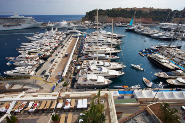A wide-angle, hyper-realistic, elevated shot of Port Hercules in Monaco, filled with hundreds of luxury yachts and superyachts. The crowded marina is surrounded by the rocky coastline, with the city and mountains in the background. In the foreground, people are relaxing in lounge chairs on a rooftop patio.