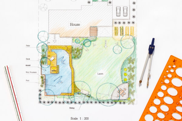 A detailed, colorful landscape blueprint and hand-drawn plan of a residential backyard garden. The design includes a swimming pool, a patio with a wall fountain, a deck, a lawn, and various plants. The document is on a white table with architectural drawing tools beside it.
