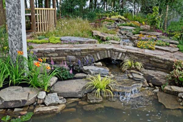 A wide-angle view of a professionally maintained backyard with a beautiful water feature. A small, handcrafted stone bridge crosses a pond with a waterfall, surrounded by diverse plants, flowers, and natural rocks.