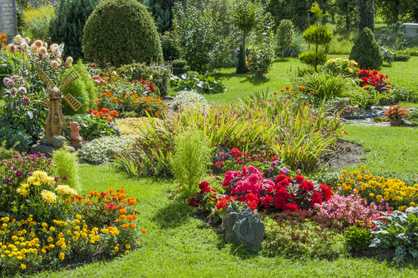 A vibrant, sunny view of a professionally maintained residential garden with a perfectly manicured lawn, lush bushes, and a variety of colorful flowers, including red, orange, pink, and yellow. A miniature windmill statue is visible in the background, adding a touch of whimsy.