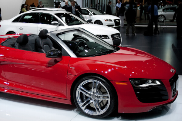 A side-angle view of a red Audi R8 Spyder convertible sports car with a V10 engine, on display at a brightly lit auto show. Another white Audi sedan is visible in the background, with people and other cars in the distance.