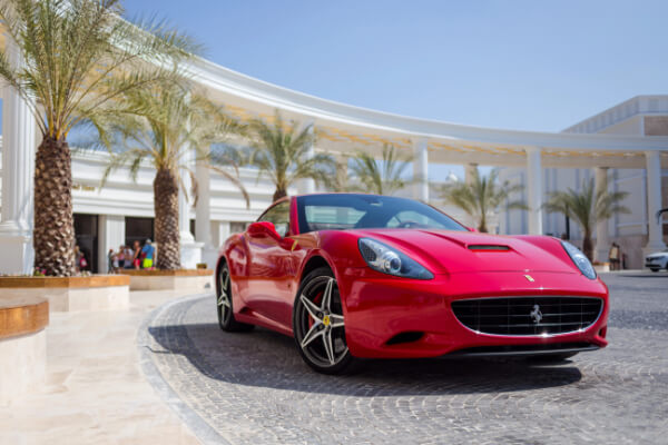 A front-angle, eye-level shot of a red Ferrari California T luxury sports car parked in front of a modern, opulent building with palm trees. The car has a shiny red finish and black alloy wheels, with a luxurious, resort-like building in the background.