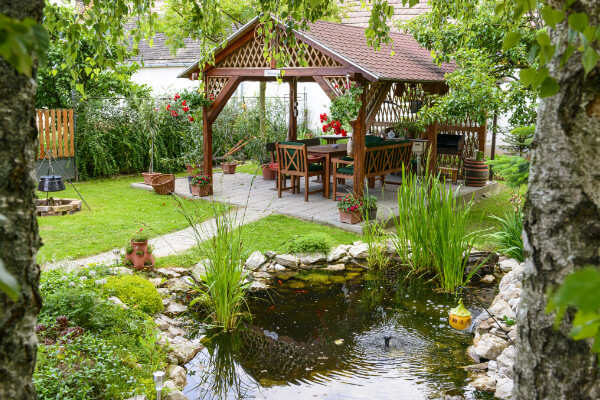 A serene, well-designed backyard garden with a lush green lawn. The space features a stone-edged pond with a small fountain and a wooden gazebo with a table, chairs, and barbecue area. The garden is surrounded by diverse plants, flowers, and trees.