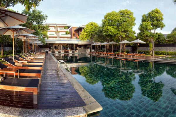 An inviting view of a large, luxurious outdoor swimming pool at a resort, surrounded by a wooden deck with rows of modern sun loungers, shade umbrellas, and lush green trees.