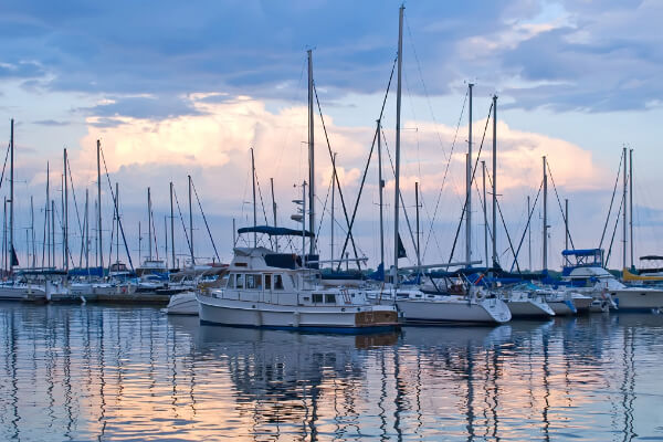 A wide-angle, low-angle shot of a marina filled with sailboats and a few powerboats docked at dusk. The calm water reflects the colorful sky and the boats' tall masts and rigging, creating a serene and peaceful scene.