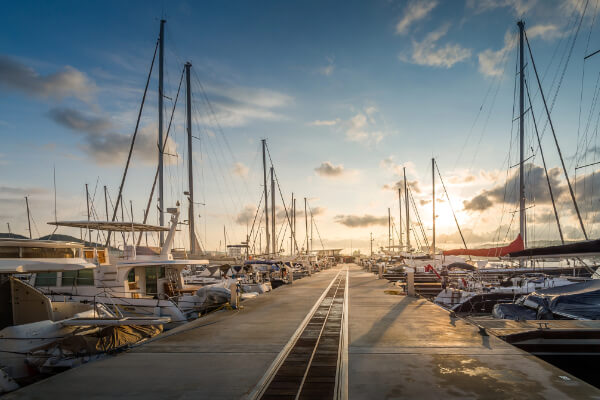 A wide-angle, eye-level shot of a marina at sunset with a long concrete pier lined on both sides with many sailboats, yachts, and catamarans. The sun is setting over the water, creating a warm golden glow and reflections on the water and pier.
