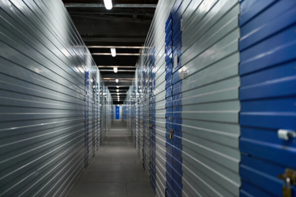 A dimly lit, slightly blurry, long hallway inside a self-storage facility, with two opposing rows of secure metal lockers. Some of the storage units have bright blue doors, while others are gray, with a lock on each one.
