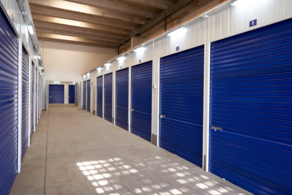 A clean, well-lit indoor storage facility hallway with two long rows of numbered, secure self-storage units with bright blue, corrugated roll-up doors and individual locks. The floor is polished concrete, and a light is visible at the end of the hall.