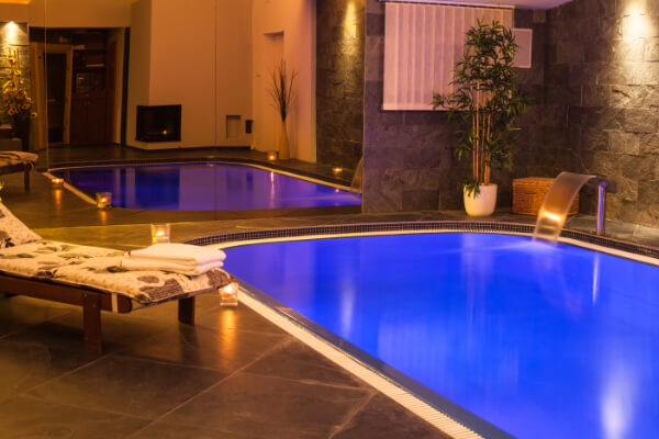 A serene indoor spa pool with warm, ambient lighting, featuring a lounge chair with fresh towels in the foreground, candles along the pool's edge, and a small waterfall jet.
