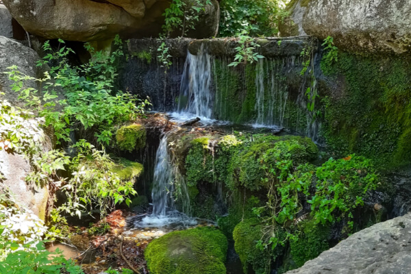 A wide-angle, hyper-realistic, eye-level shot of a small, tranquil waterfall with water cascading over natural moss-covered rocks and green foliage.