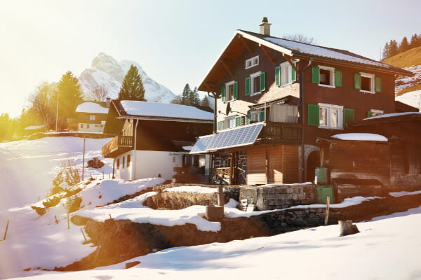 A picturesque, sun-drenched image of a traditional wooden chalet with a solar panel on its balcony, nestled in a snowy mountain landscape. The property is surrounded by snow, with a steep-sloped mountain and a bright sun flare in the background.