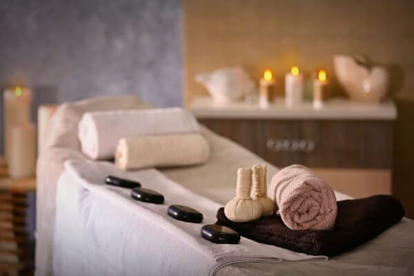 A relaxing spa scene with a massage table prepared with clean towels, a row of smooth hot stones, and herbal compress balls, with lit candles and soft lighting in the background.