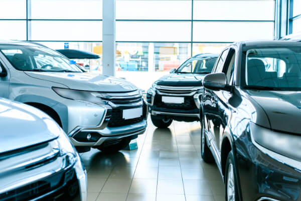 A wide-angle, hyper-realistic, and brightly lit view from inside a modern automotive group showroom. Multiple new, reflective SUVs are visible, including a gray one, a silver one, and a black one, on a polished tiled floor with large windows in the background.