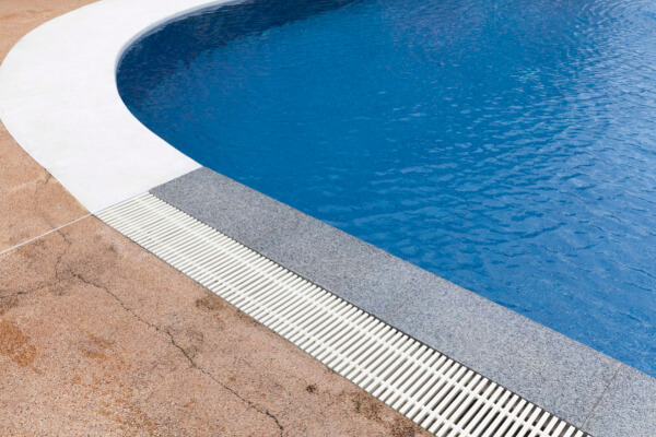 A close-up, low-angle shot of the rounded edge of a light-blue swimming pool, showing the white coping and a white plastic grate covering the perimeter overflow skimmer drain. The deck is made of brown and gray concrete.