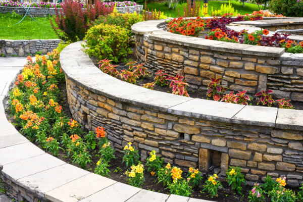 A vibrant, colorful public or residential garden with tiered beds featuring natural stone retaining walls and diverse plants and flowers. The design includes various flowers in shades of yellow, orange, and red, along with lush green foliage.