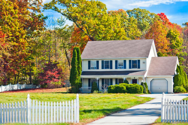 A two-story custom-built house with light-colored siding, a covered front porch, a two-car garage, and dark blue shutters on the windows. The house is surrounded by a lush green lawn, a white picket fence, and a vibrant forest with fall foliage.