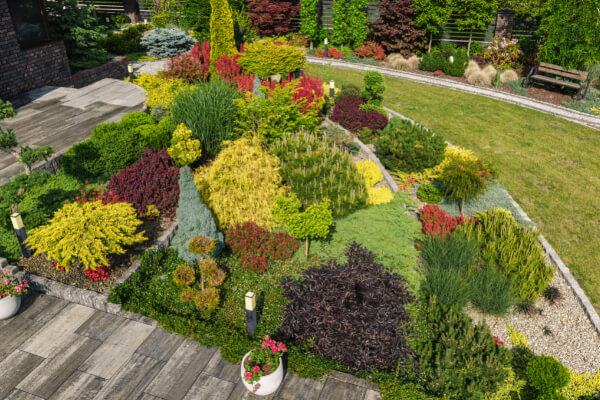 A vibrant, sunny view of a professionally maintained residential garden with a perfectly manicured lawn, lush bushes, and a variety of colorful flowers, including red, orange, pink, and yellow. A miniature windmill statue is visible in the background, adding a touch of whimsy.