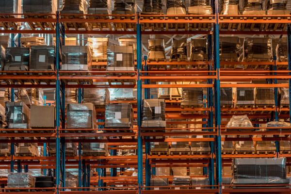 A wide-angle, hyper-realistic, and low-angle shot of tall warehouse racks filled with a variety of goods on wooden pallets. The shelves are packed with inventory, including shrink-wrapped boxes, with a dark and moody lighting effect.