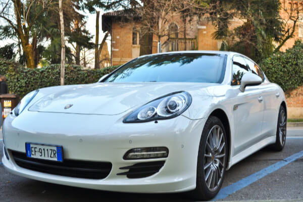 A front-angle, eye-level shot of a parked white Porsche Panamera luxury car with a shiny finish. The car is on a paved street with a building and lush green bushes in the background, showing a European license plate.
