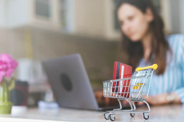 A close-up, high-angle shot of a miniature metal shopping cart with red and white credit cards inside. A young woman is sitting in the background, out of focus, using a laptop, representing the concept of e-commerce and online payments.