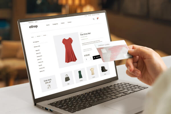A close-up, high-angle shot of a woman's hand holding a silver credit card over a laptop keyboard. The laptop screen shows an e-commerce website with a red dress, bags, and shoes on a shopping page. The scene is brightly lit with a blurry background.