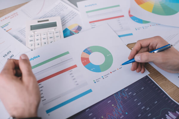 Hands using a blue pen to point at an analytics report with a colorful donut chart, surrounded by a calculator and various data printouts.
