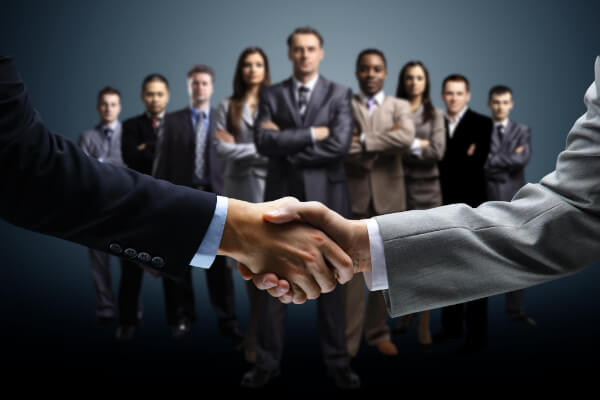 A close-up handshake in the foreground with a diverse, professional sales team standing confidently in the background.