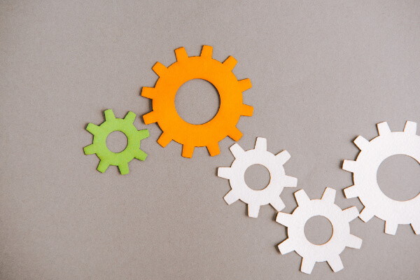 Four interlocking paper cutout gears in orange, green, and white colors placed on a neutral grey background.