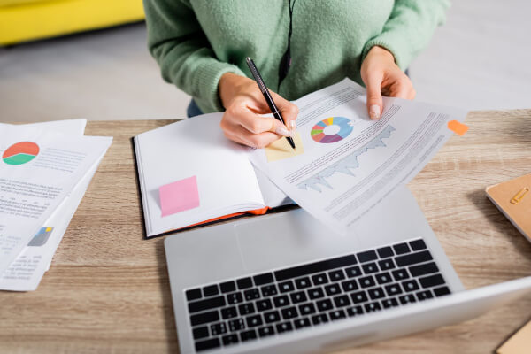 A person in a green sweater writing in a notebook while reviewing AI-generated charts and graphs on a desk next to a laptop.