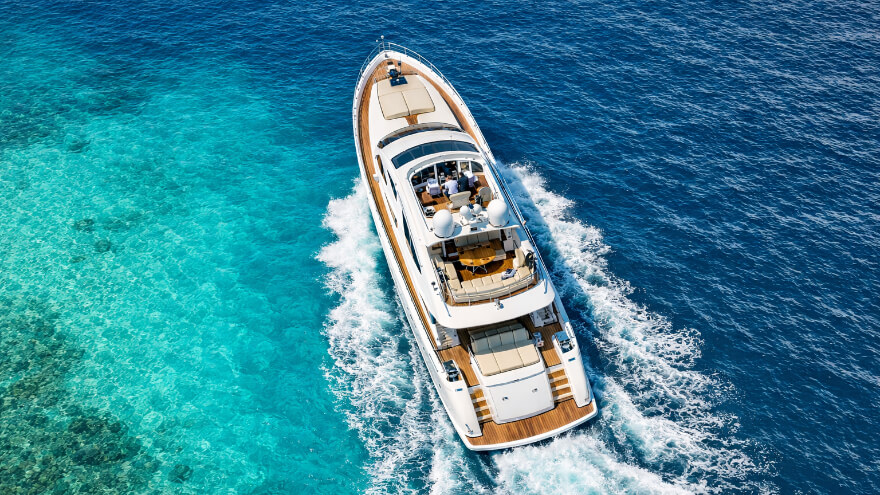 Overhead view of a sleek white luxury motor yacht sailing through clear turquoise ocean water with visible sun loungers and dining area.
