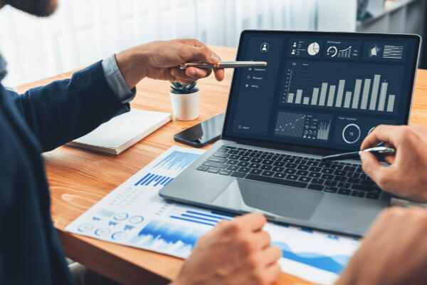 Two professionals pointing at a laptop screen displaying an AI marketing dashboard with bar charts, gauges, and real-time performance metrics.