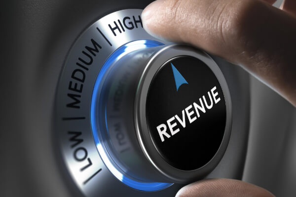 A hand turning a metallic dial labeled "REVENUE" from medium to high, symbolizing the impact of AI on sales performance metrics.
