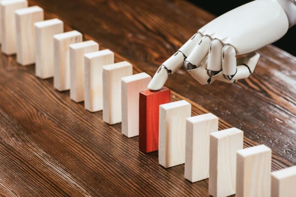 A white robotic finger touching a red wooden block in a line of standing dominoes to trigger a chain reaction.