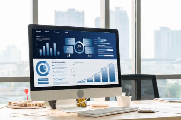 A desktop computer monitor sitting on a bright office desk, displaying a dark blue financial analytics interface with bar graphs and pie charts.