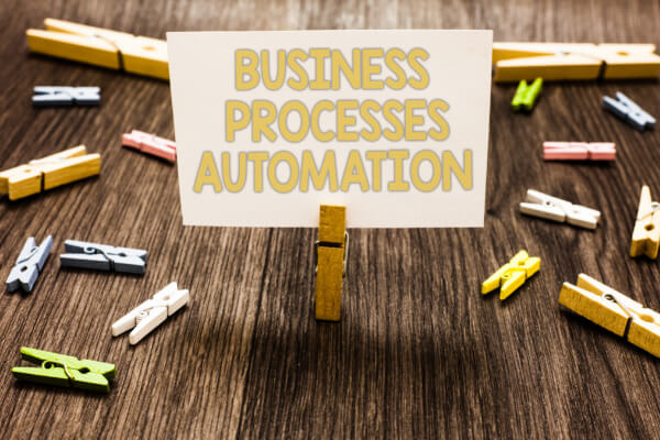 A white paper card displaying the text BUSINESS PROCESSES AUTOMATION held upright by a wooden clothespin on a dark wooden surface scattered with colorful clothespins.