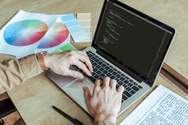 Hands typing on a laptop displaying code, situated on a wooden desk next to a printed color wheel and palette swatches.