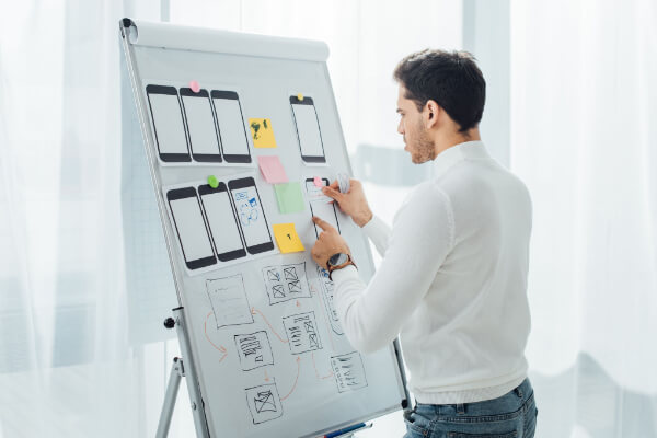 A man standing at a whiteboard, moving sticky notes around printed mobile phone screen wireframes.