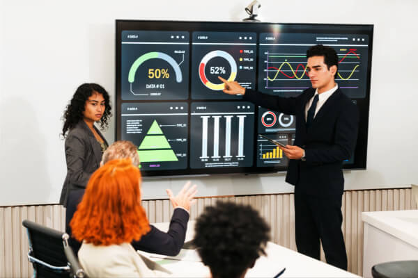 A man presenting a dark-themed business analytics dashboard on a large digital screen to a team of professionals.